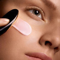 Primo piano di una donna che applica una crema rosa sulla guancia con un applicatore nero e oro.