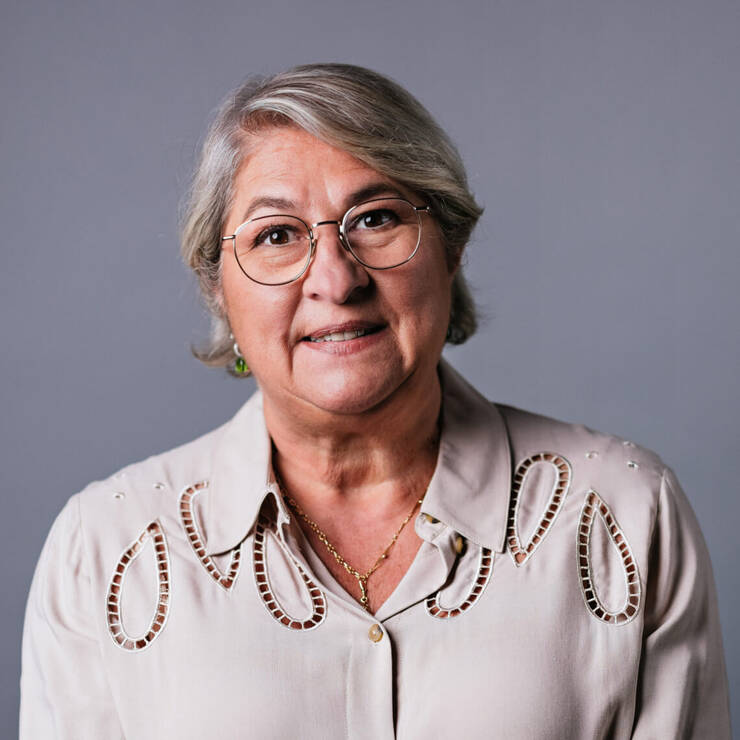 Donna di mezza eta con occhiali, sorridente, indossa una camicia beige con dettagli in pizzo, su sfondo grigio.
