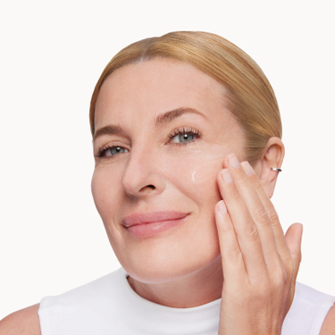 Donna sorridente che applica una crema viso trasparente sulla guancia, pelle matura e luminosa