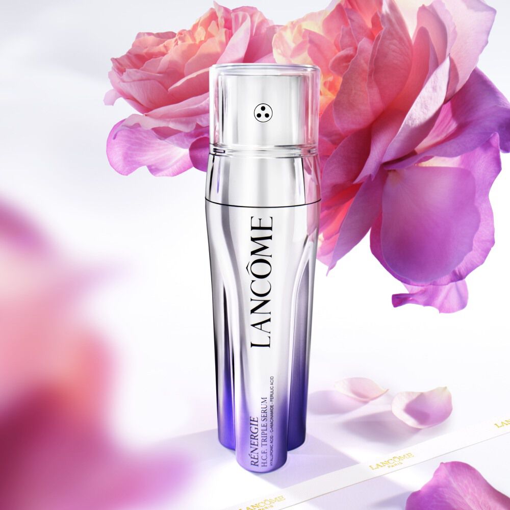 Una boccetta di siero della linea Renergie di Lancome, circondata da petali di rosa su uno sfondo luminoso.
