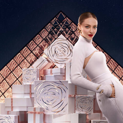 Una donna elegante con abito bianco posa accanto a torri di scatole regalo bianche, con il Louvre sullo sfondo illuminato dalla notte.