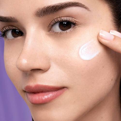 Una giovane donna con pelle luminosa applica una crema sul viso, sorridendo e guardando verso l'obiettivo.