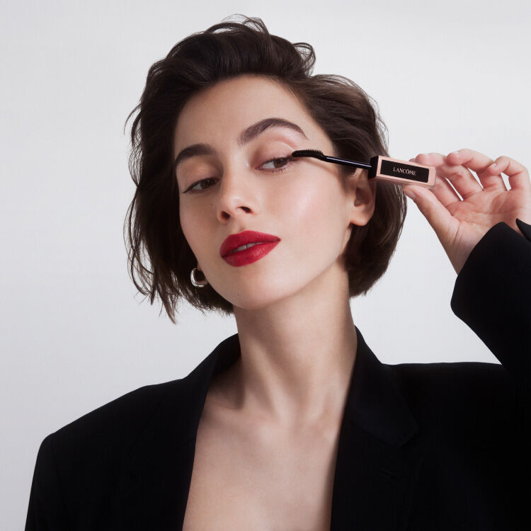 Una donna con capelli corti e scuri applica un mascara, indossando un blazer nero e un rossetto rosso.