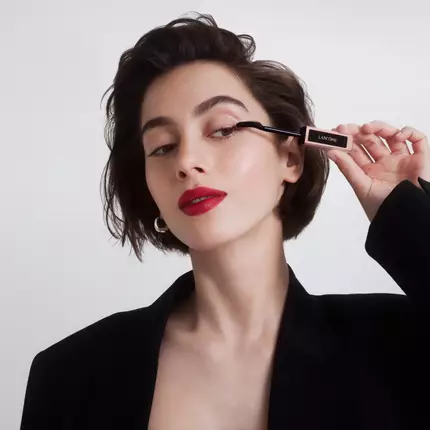 Una donna con capelli corti e scuri applica un mascara, indossando un blazer nero e un rossetto rosso.
