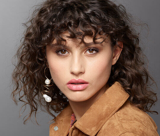 Immagine ravvicinata di una giovane donna con capelli ricci e scuri, indossando orecchini eleganti e un giubbotto di camoscio. Ha un trucco naturale e uno sguardo intenso.