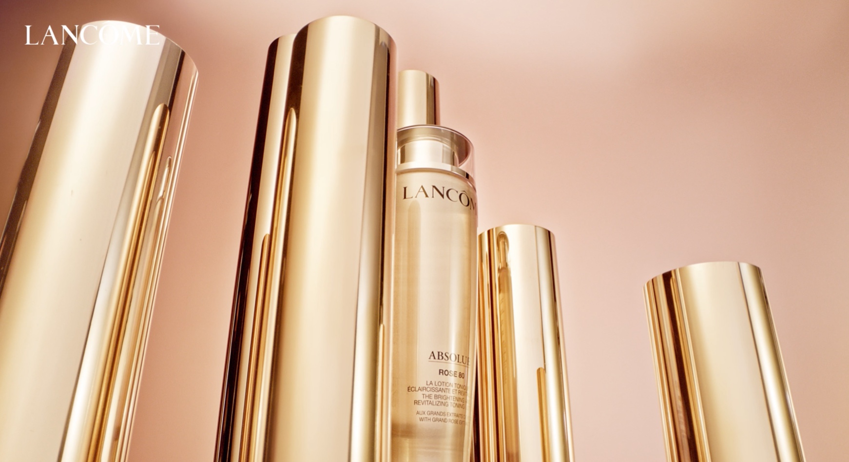 Flaconi di prodotti Lancôme in oro lucido, disposti elegantemente su uno sfondo rosa chiaro.